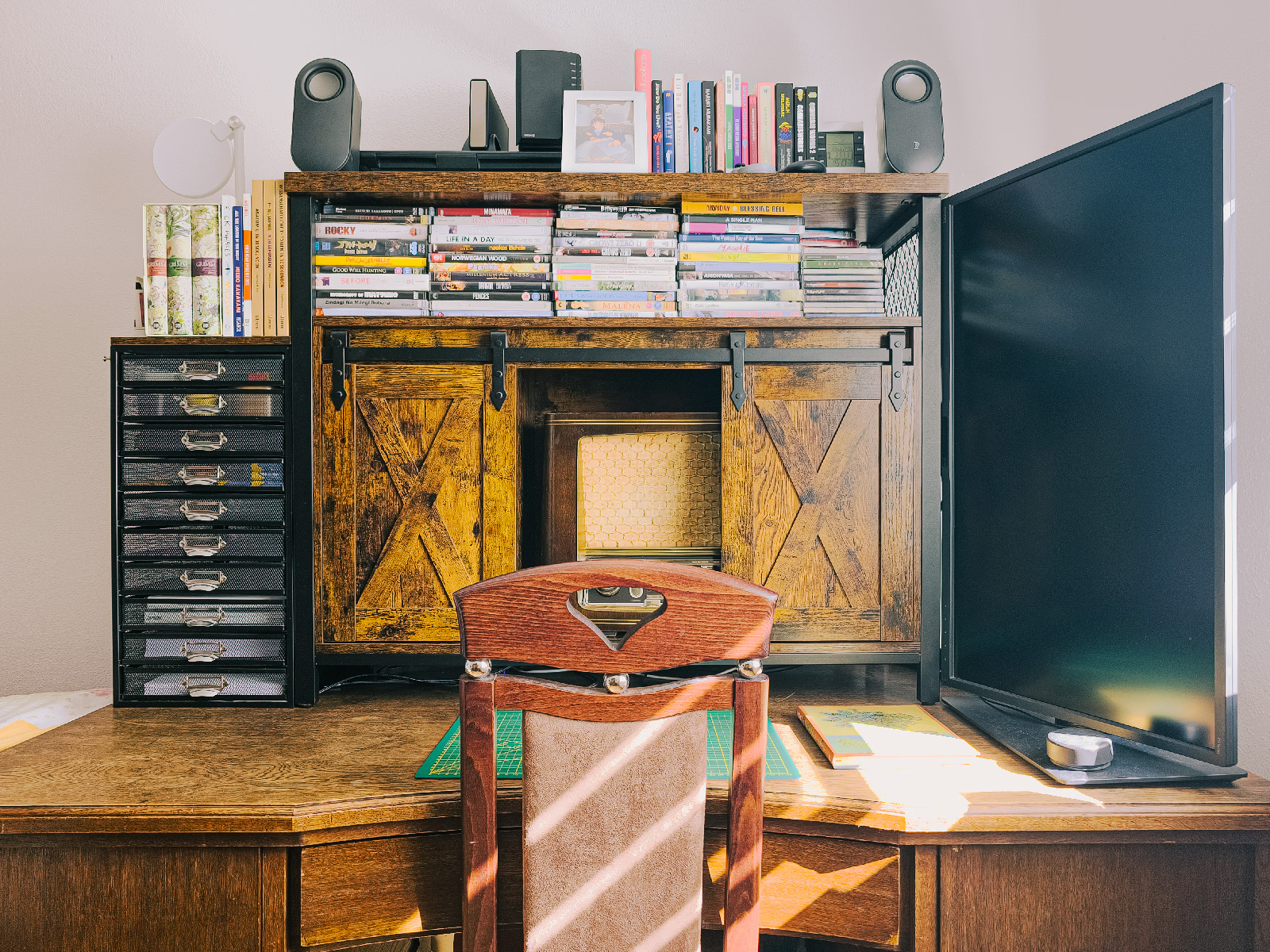 Wooden chair in front of an old hardwood desk with a vertical monitor, a drawer containing a vintage radio, and stacks of DVDs, books, and small electronic devices.